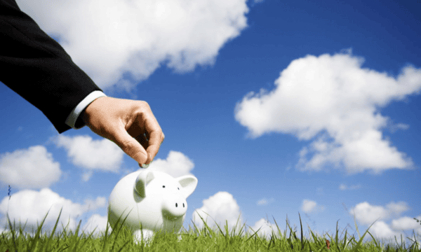 Male hand placing coin in piggybank with blue sky, clouds, and grass