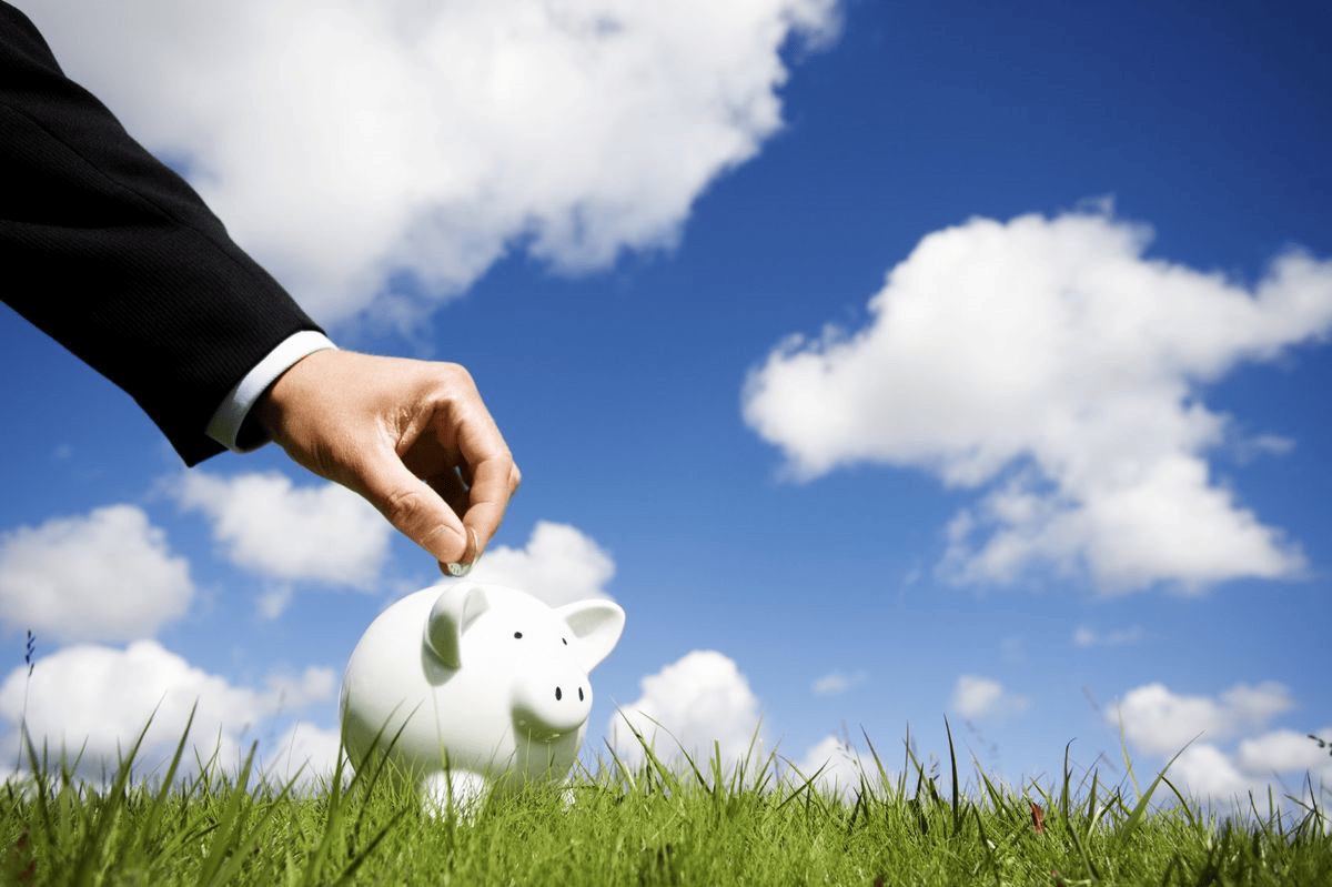 Male hand placing coin in piggybank with blue sky, clouds, and grass